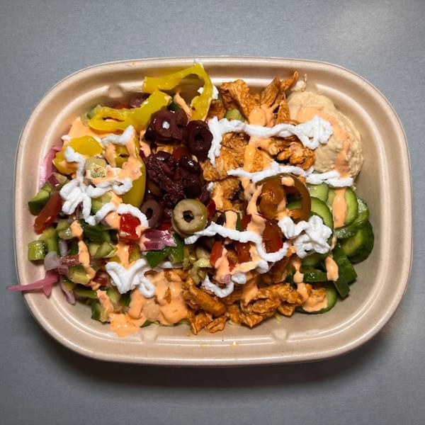 A Build-A-Pita & Pizza bowl with chicken, hummus, olives, peppers, and various vegetables, drizzled with sauces.