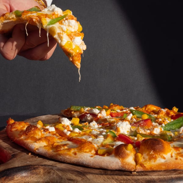 Hand lifting a cheesy slice of Build-A-Pita & Pizza's vegetable pizza from a wooden board.