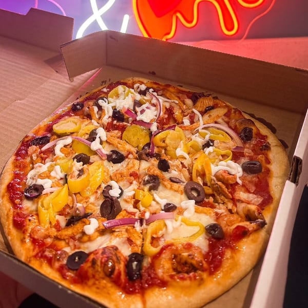 A Build-A-Pita & Pizza Restaurant pizza in a box, topped with chicken, olives, onions, and peppers.
